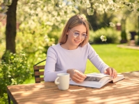 A cute young teenage girl is reading a book while sitting at a wooden table with a Cup of coffee, in the back yard.