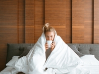 Cute teen girl under blanket in white bed in bedroom woken up and drink tea with laptop. Breakfast in bed, lazy morning